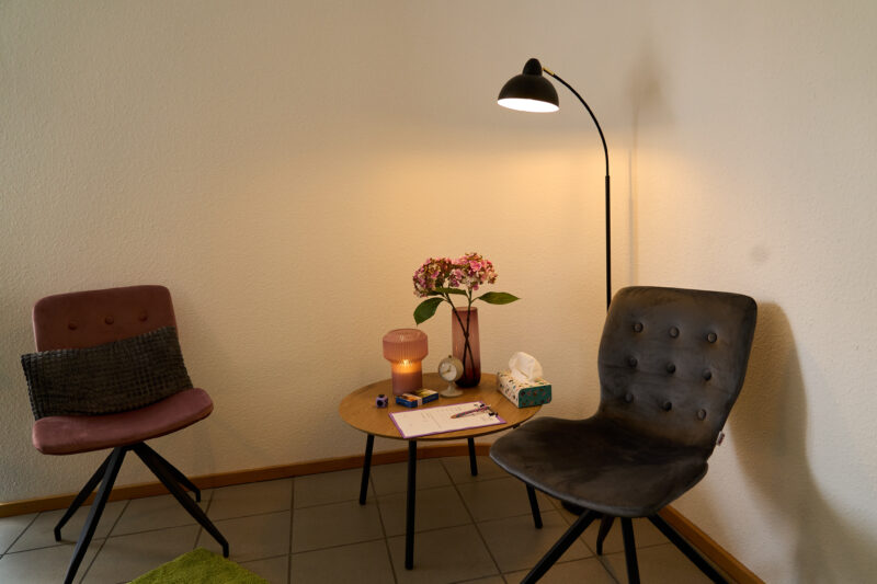 Ein Ausschnitt der Beratungsecke mit zwei gepolsterten Stühlen, einem weichen Teppich, einer Stehlampe und einem kleinen Tisch, auf dem ein paar Utensilien bereitstehen.