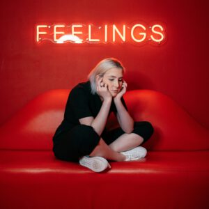 Eine junge Frau sitzt nachdenklich auf einem roten Sofa unter einem leuchtenden Schild mit dem Schriftzug "Feelings"
