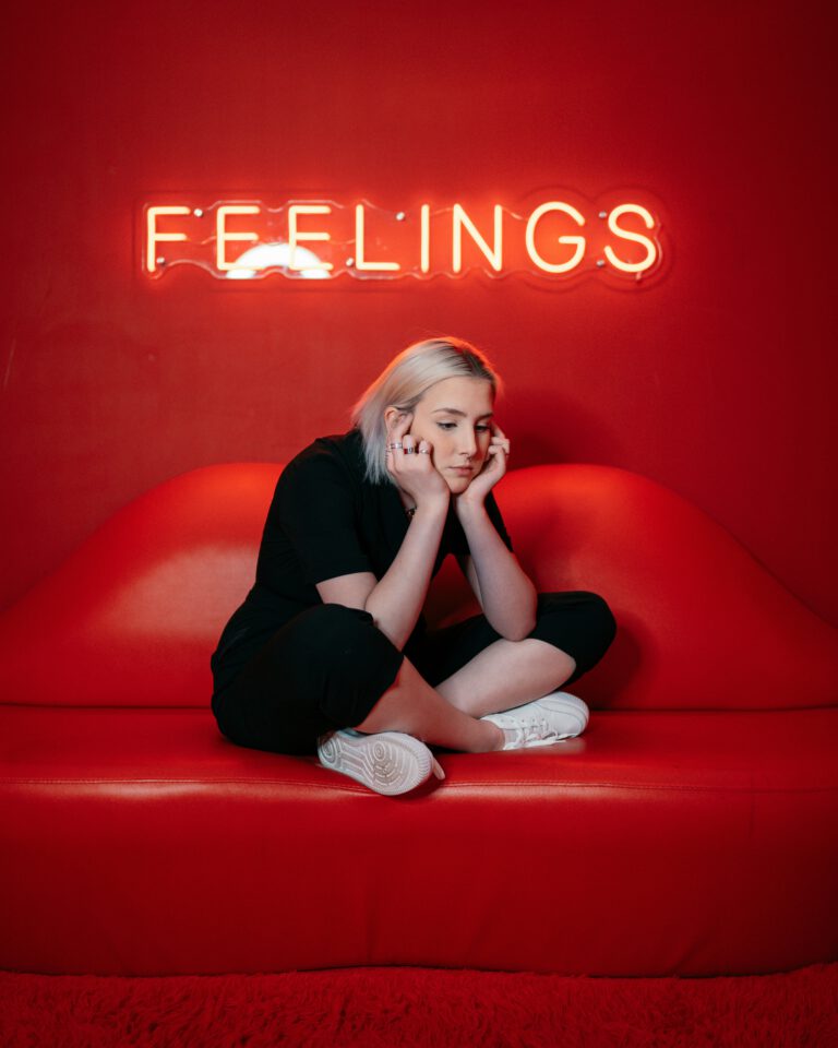 Eine junge Frau sitzt nachdenklich auf einem roten Sofa unter einem leuchtenden Schild mit dem Schriftzug "Feelings"