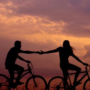 Die Silhouetten von zwei Menschen auf Fahrrädern, die sich vor der Abenddämmerung die Hand reichen.