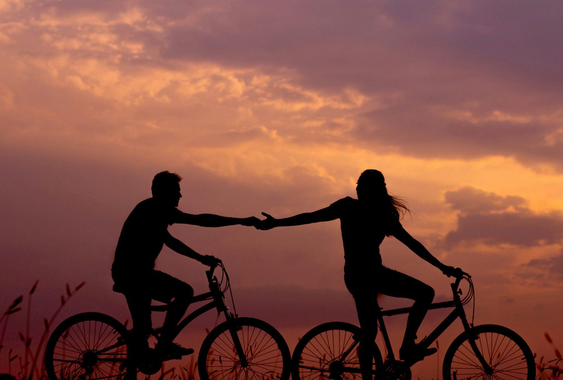 Die Silhouetten von zwei Menschen auf Fahrrädern, die sich vor der Abenddämmerung die Hand reichen.